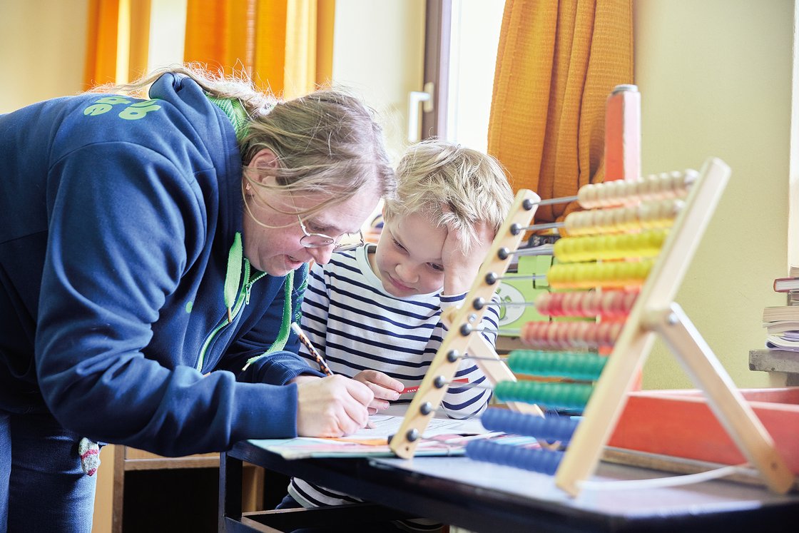 Eine Frau hilft einem kleinen Jungen bei den Schularbeiten an einem Tisch. Ein Abakus mit bunten Perlen ist im Vordergrund.