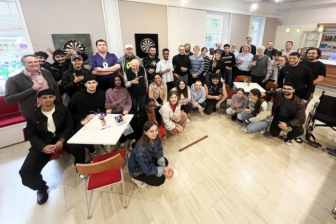 Eine vielfältige Gruppe Menschen jeden Alters lachend in einem hellen Raum versammelt, einige mit Getränken, Dartscheiben an der Wand.