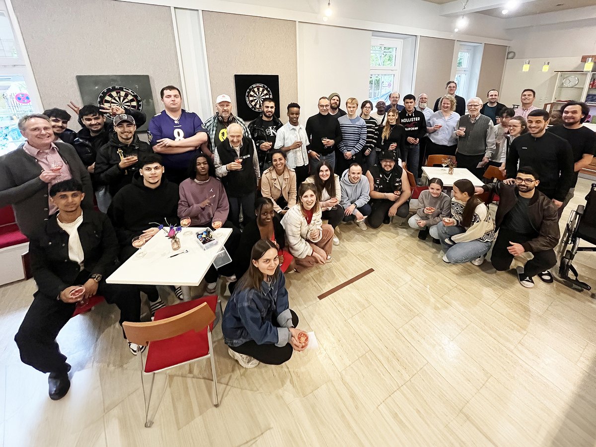 Eine vielfältige Gruppe Menschen jeden Alters lachend in einem hellen Raum versammelt, einige mit Getränken, Dartscheiben an der Wand.