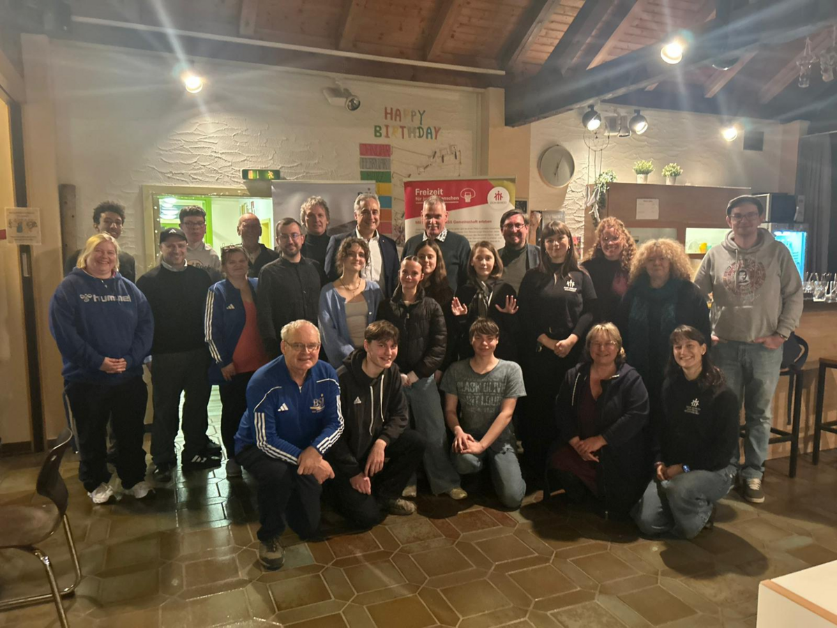 Eine große, vielfältige Gruppe von Erwachsenen und Jugendlichen lächelt für ein Gruppenfoto in Innenräumen.