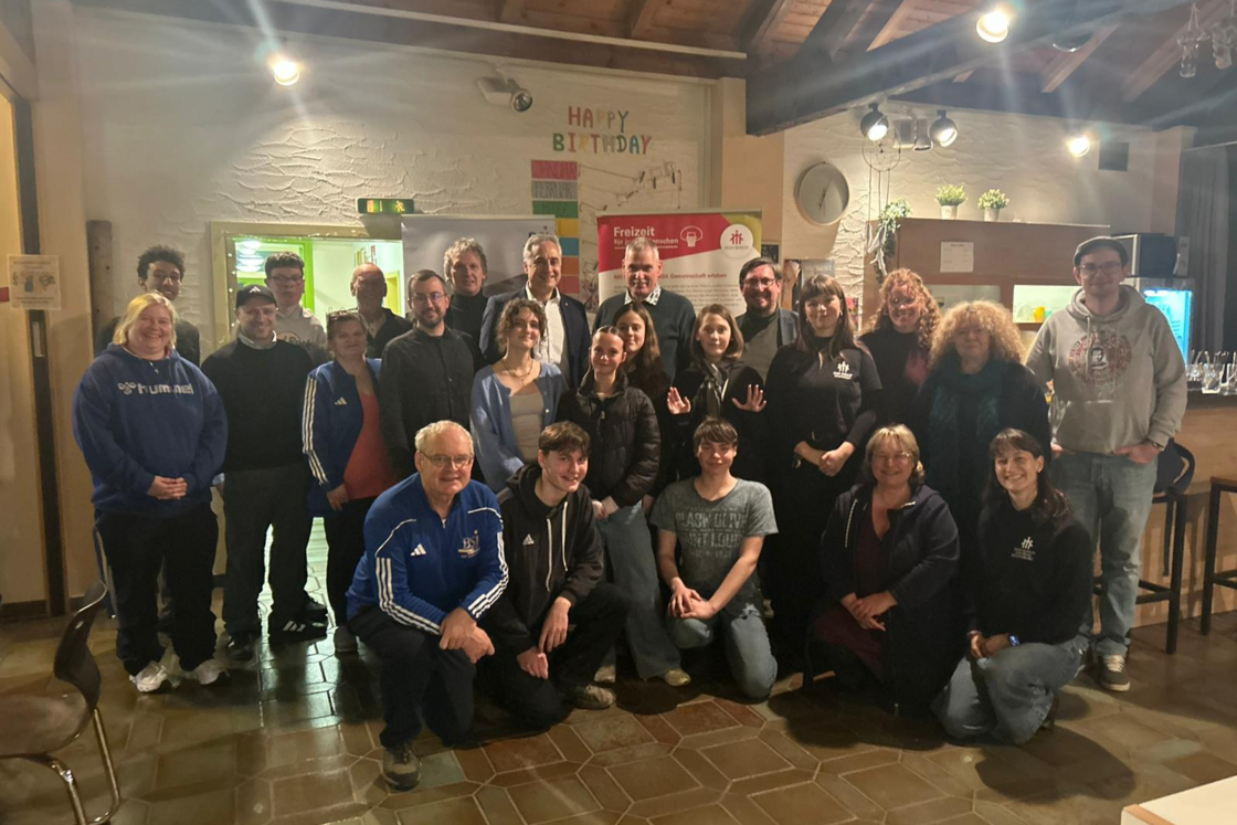 Eine große, vielfältige Gruppe von Erwachsenen und Jugendlichen lächelt für ein Gruppenfoto in Innenräumen.