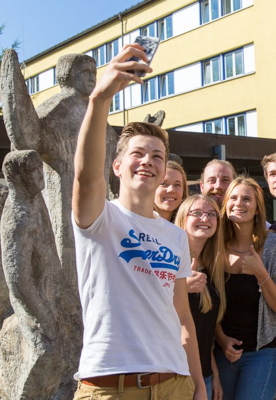 Gruppe lächelnder Studierender macht ein Selfie vor einem gelb-apricotfarbenen Gebäude und einer Skulptur.