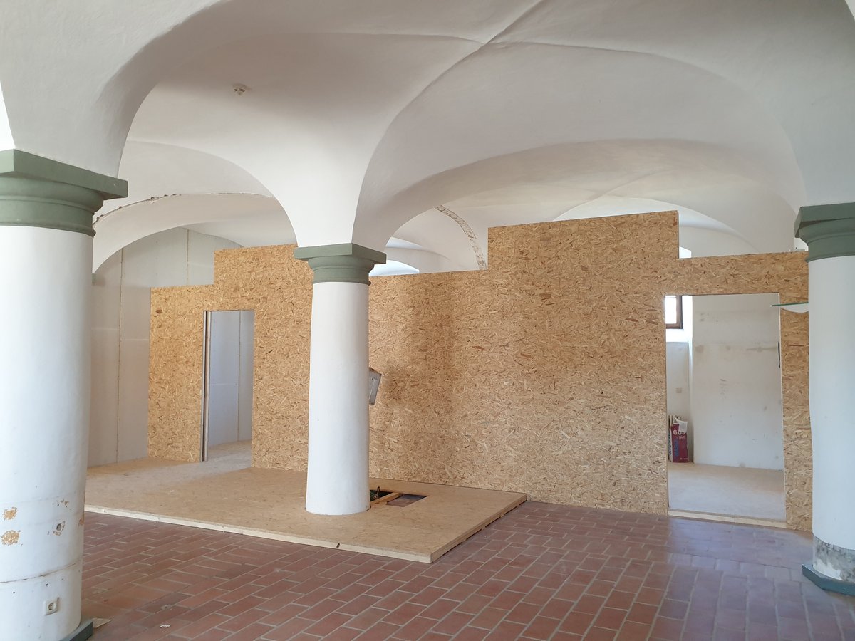 Halle mit weißen Gewölbedecken und Säulen, die durch temporäre Spanplattenwände und Holzböden unterteilt ist.