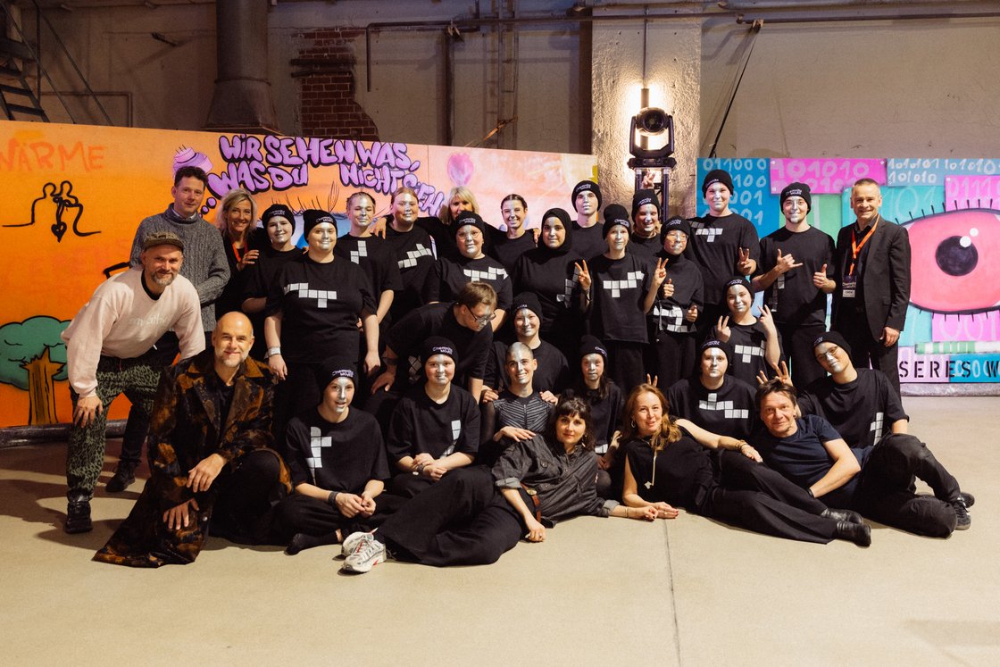 Gruppenfoto von rund 30 Personen in schwarzen Shirts, einige mit weißer Gesichtsbemalung, vor einer bunten Graffitiwand.