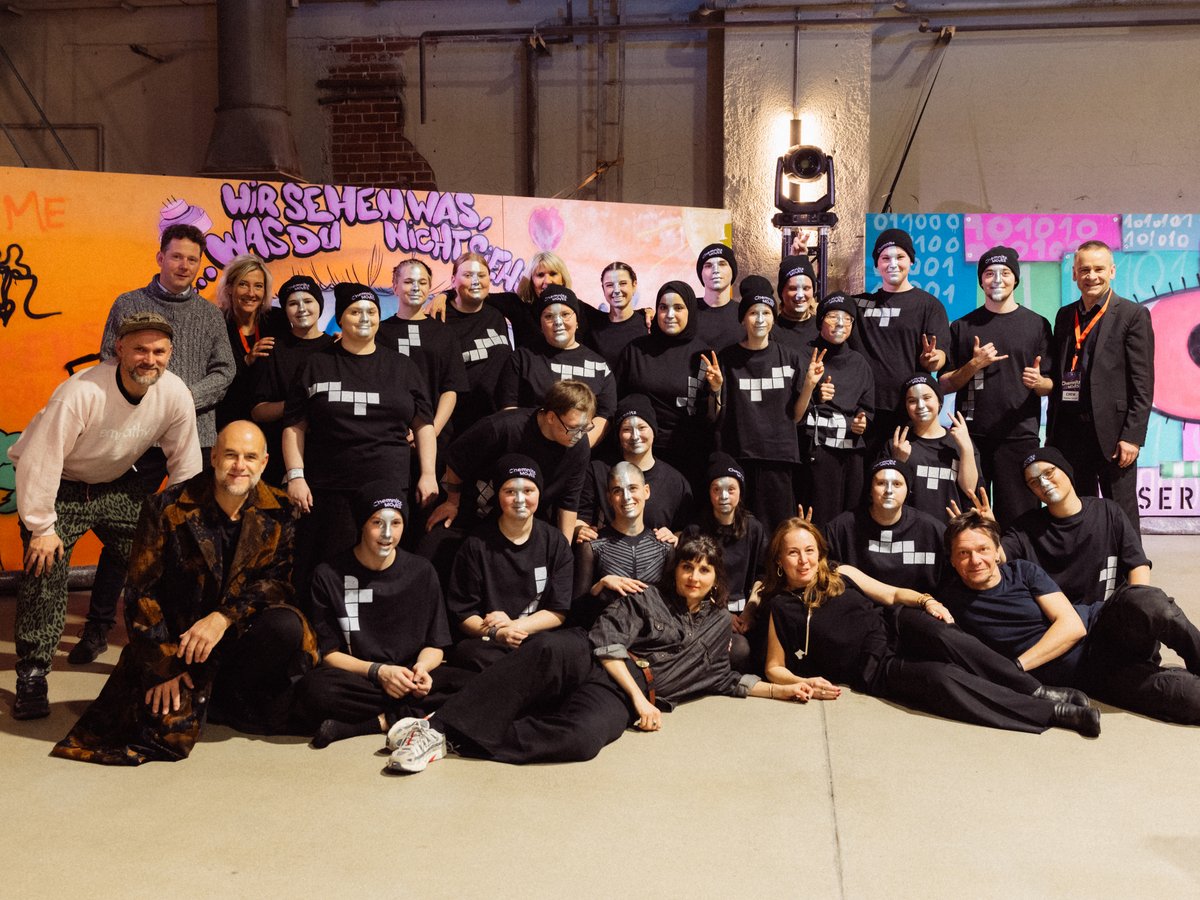 Gruppenfoto von rund 30 Personen in schwarzen Shirts, einige mit weißer Gesichtsbemalung, vor einer bunten Graffitiwand.