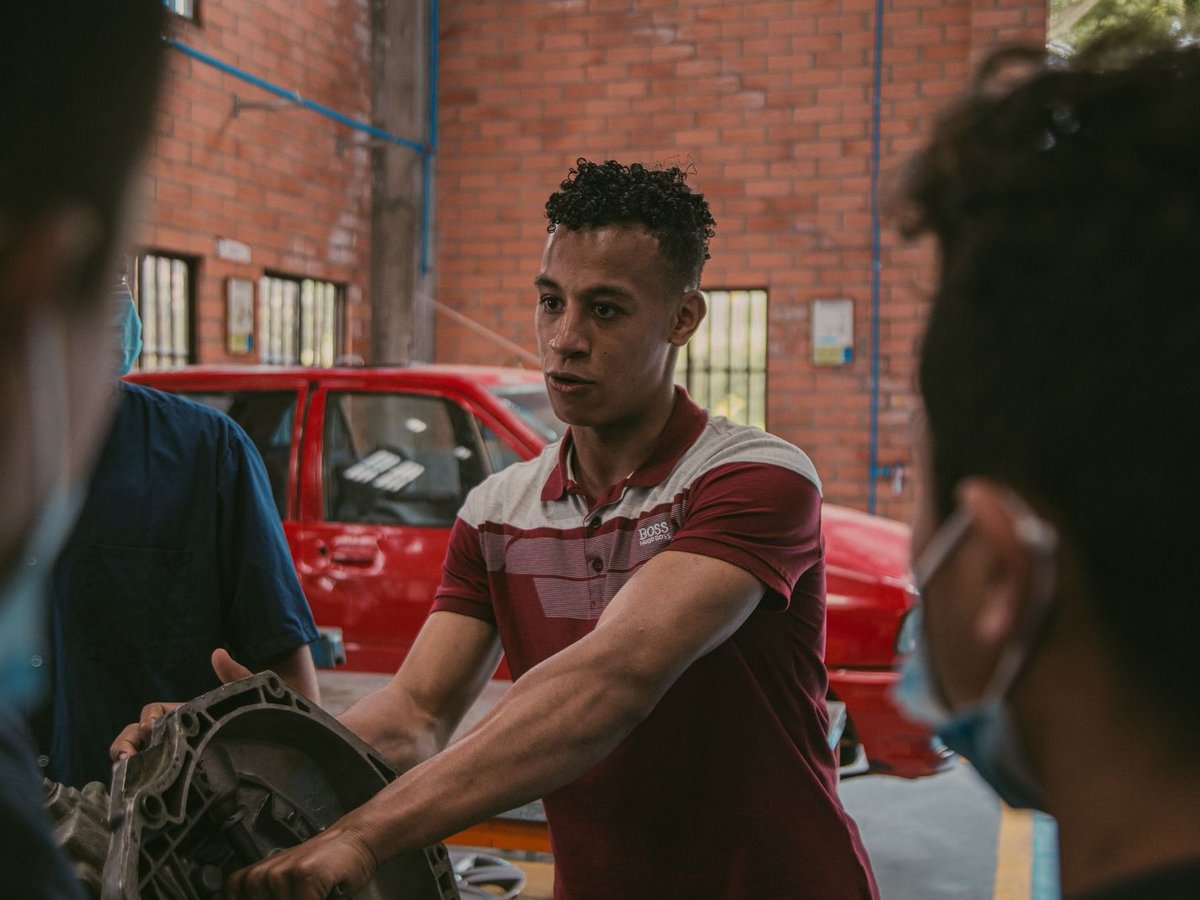 Junger Mann erklärt Autogetriebe in Kfz-Werkstatt vor anderen Studenten.