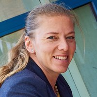 Eine lächelnde Frau mit blonden Haaren in einem dunkelblauen Blazer, hellblauen Augen und einem hellblauen Ohrring. Verschwommener blau-grüner Hintergrund.