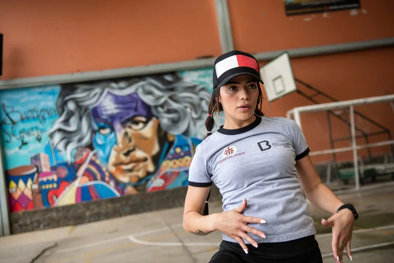 Junge Frau in Kappe und grauem T-Shirt tanzt auf einem Sportplatz, ein lebendiges Beethoven-Wandgemälde im Hintergrund.