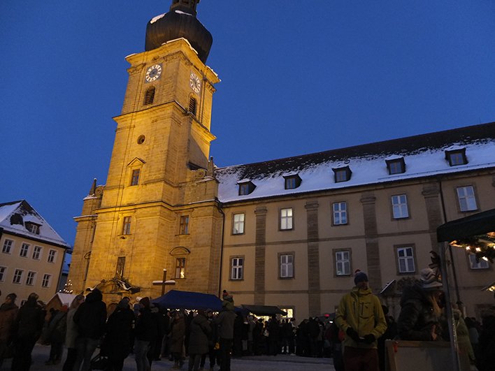 Weihnachtsmarkt bei Nacht vor einer historischen Kirche mit Glockenturm und verschneiten Dächern, warm beleuchtet.