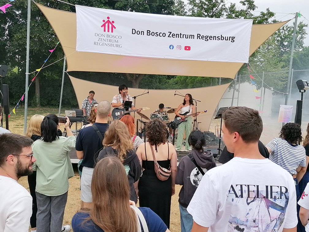 Eine Band spielt ein Open-Air-Konzert im Don Bosco Zentrum Regensburg. Sänger, Gitarristin, Schlagzeuger und Keyboarder treten vor Publikum auf.