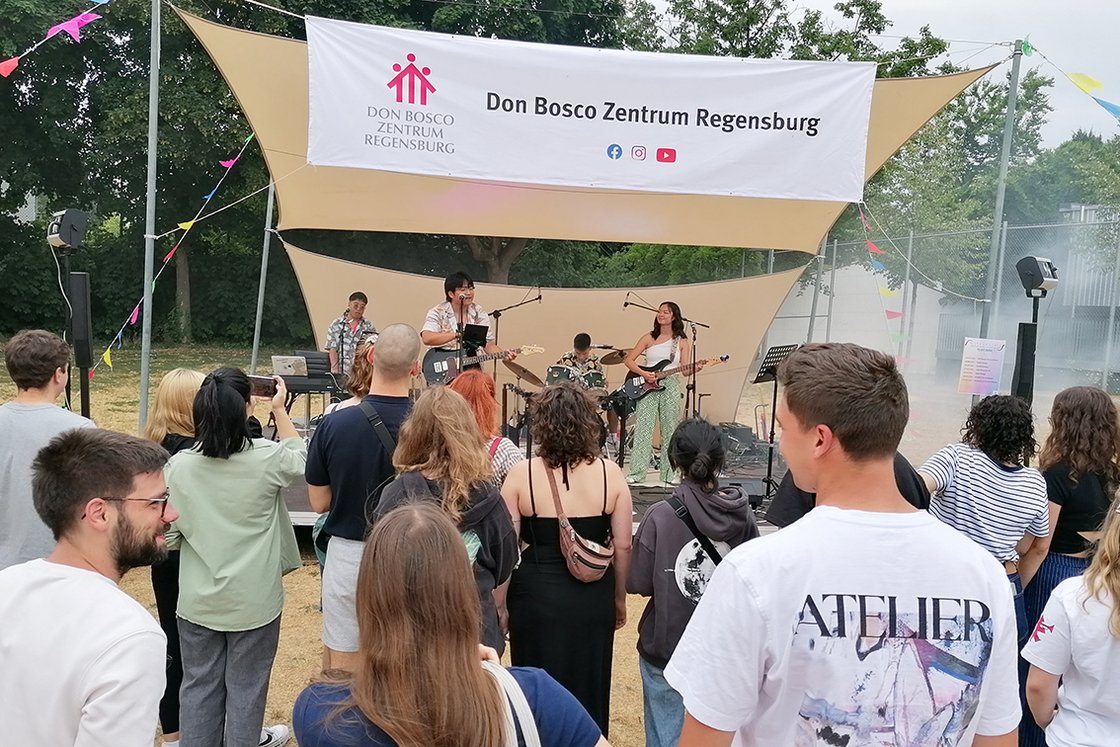 Eine Band spielt ein Open-Air-Konzert im Don Bosco Zentrum Regensburg. Sänger, Gitarristin, Schlagzeuger und Keyboarder treten vor Publikum auf.