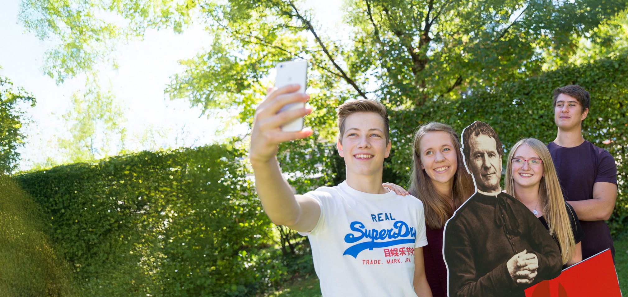 Jugend-Selfie mit Pappaufsteller Fünf fröhliche Jugendliche, darunter ein Pappaufsteller eines Priesters, machen ein Selfie im sonnigen Park.