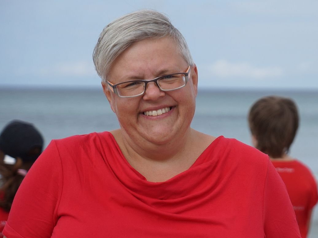 Lächelnde Frau mit grauen Haaren und Brille in rotem T-Shirt, unscharfe Kinder in roten Shirts blicken aufs Meer.