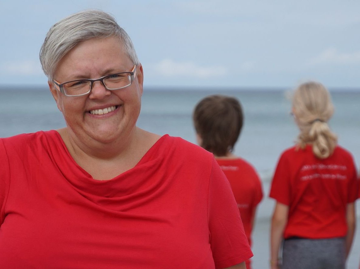 Lächelnde Frau mit grauen Haaren und Brille in rotem T-Shirt, unscharfe Kinder in roten Shirts blicken aufs Meer.