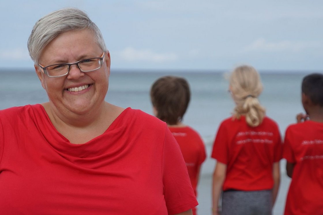 Lächelnde Frau mit grauen Haaren und Brille in rotem T-Shirt, unscharfe Kinder in roten Shirts blicken aufs Meer.