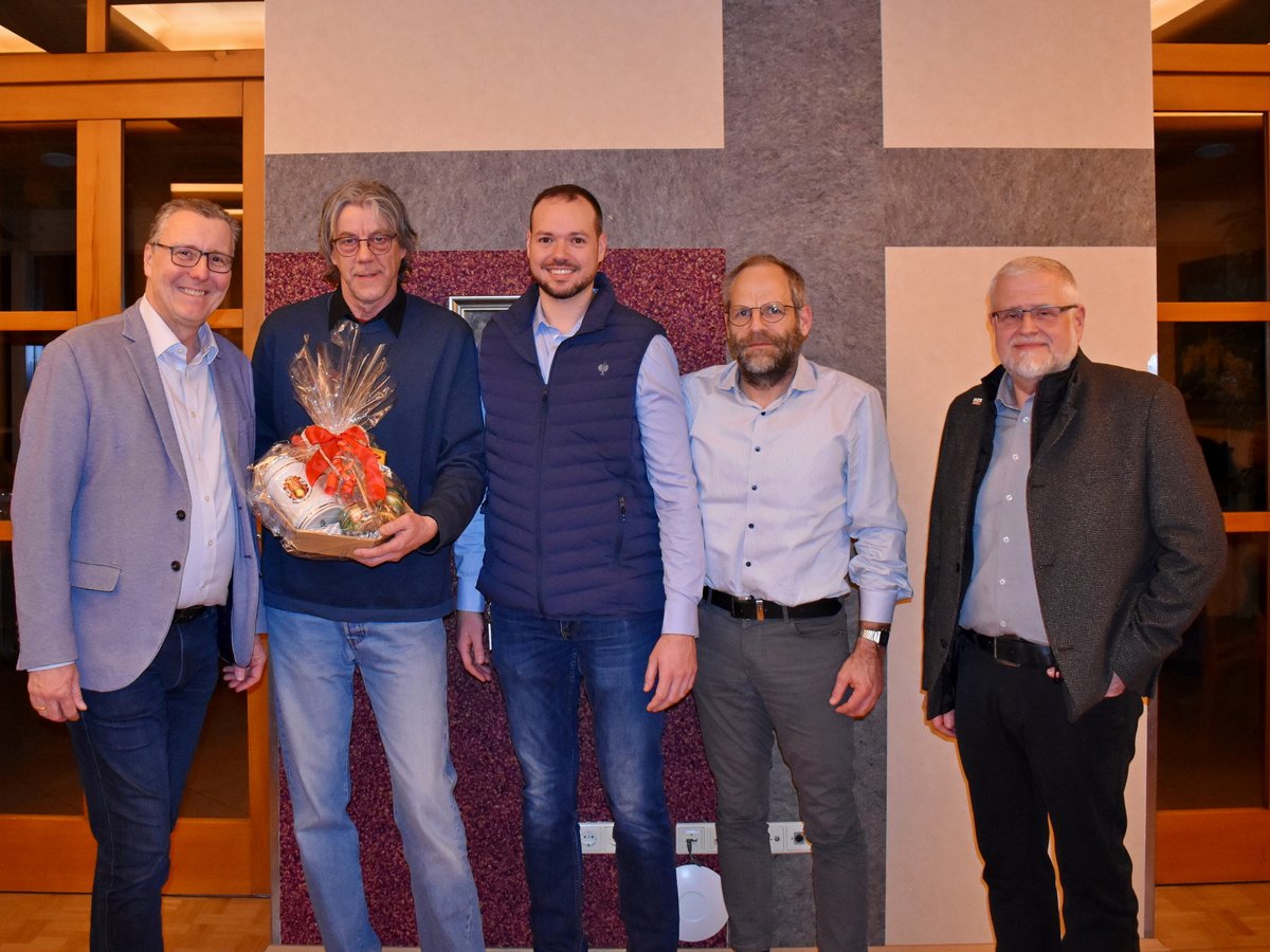 Fünf lächelnde Männer posieren für ein Foto. Der zweite Mann von links hält einen Geschenkkorb mit roter Schleife. Innenaufnahme.