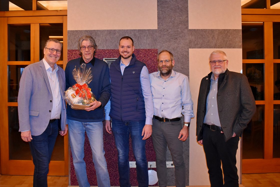 Fünf lächelnde Männer posieren für ein Foto. Der zweite Mann von links hält einen Geschenkkorb mit roter Schleife. Innenaufnahme.