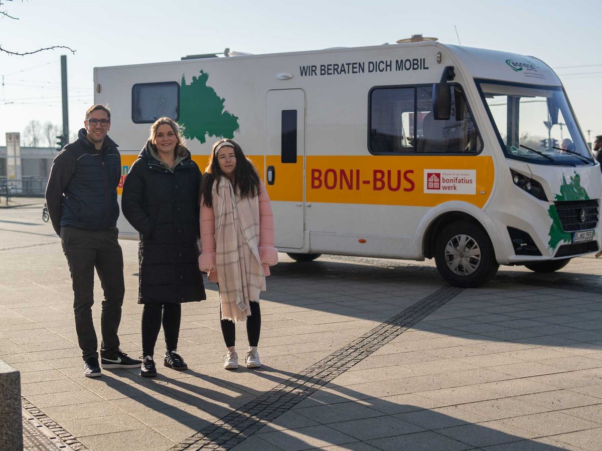 Ein Mann und zwei Frauen stehen vor einem weißen Boni-Bus mit der Aufschrift „Wir beraten dich mobil“ auf einem sonnigen Platz.