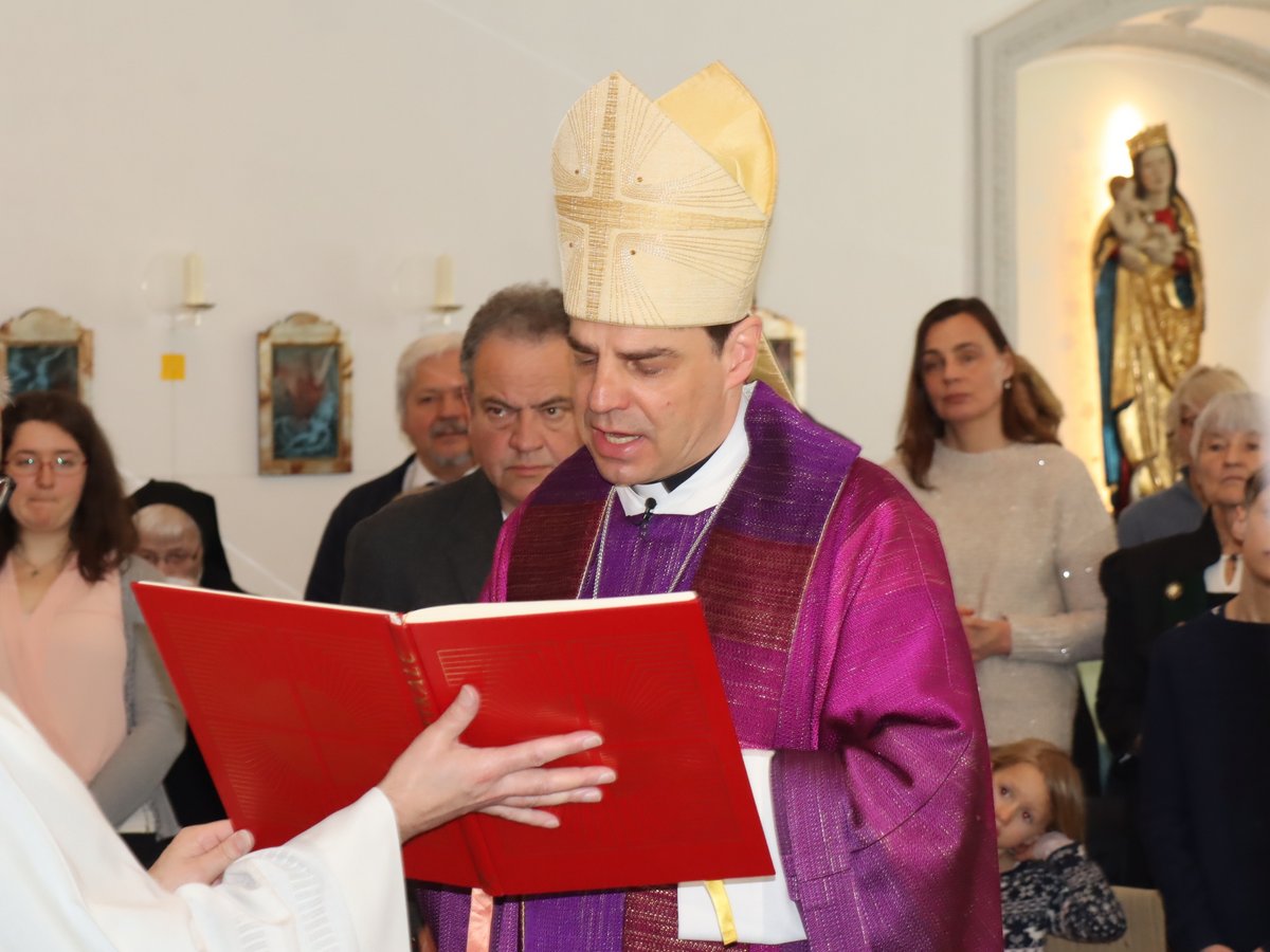 Bischof in Mitra und violettem Gewand liest aus einem roten Buch während eines Gottesdienstes, Zuschauer beobachten.