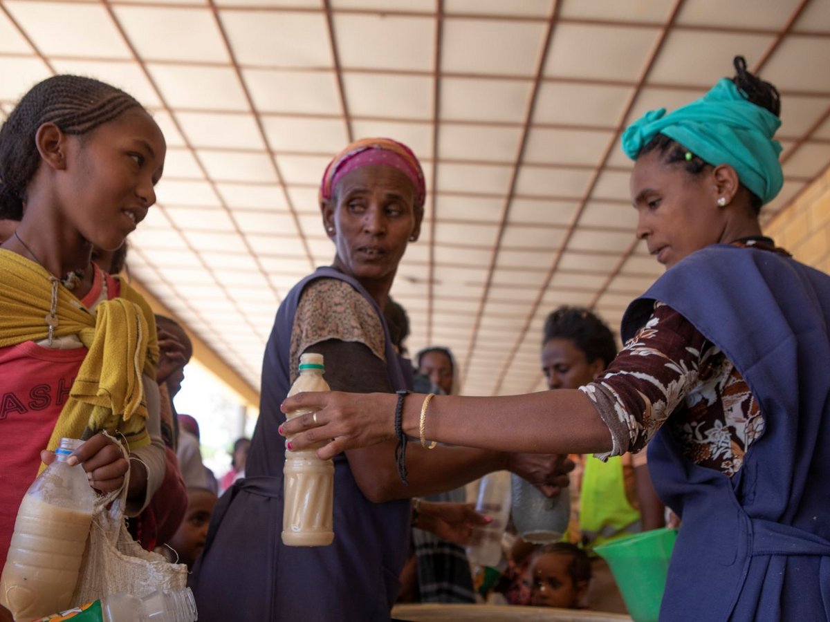 Frauen und Kinder unter einem Dach. Eine Frau reicht einer anderen eine Flasche mit heller Flüssigkeit.