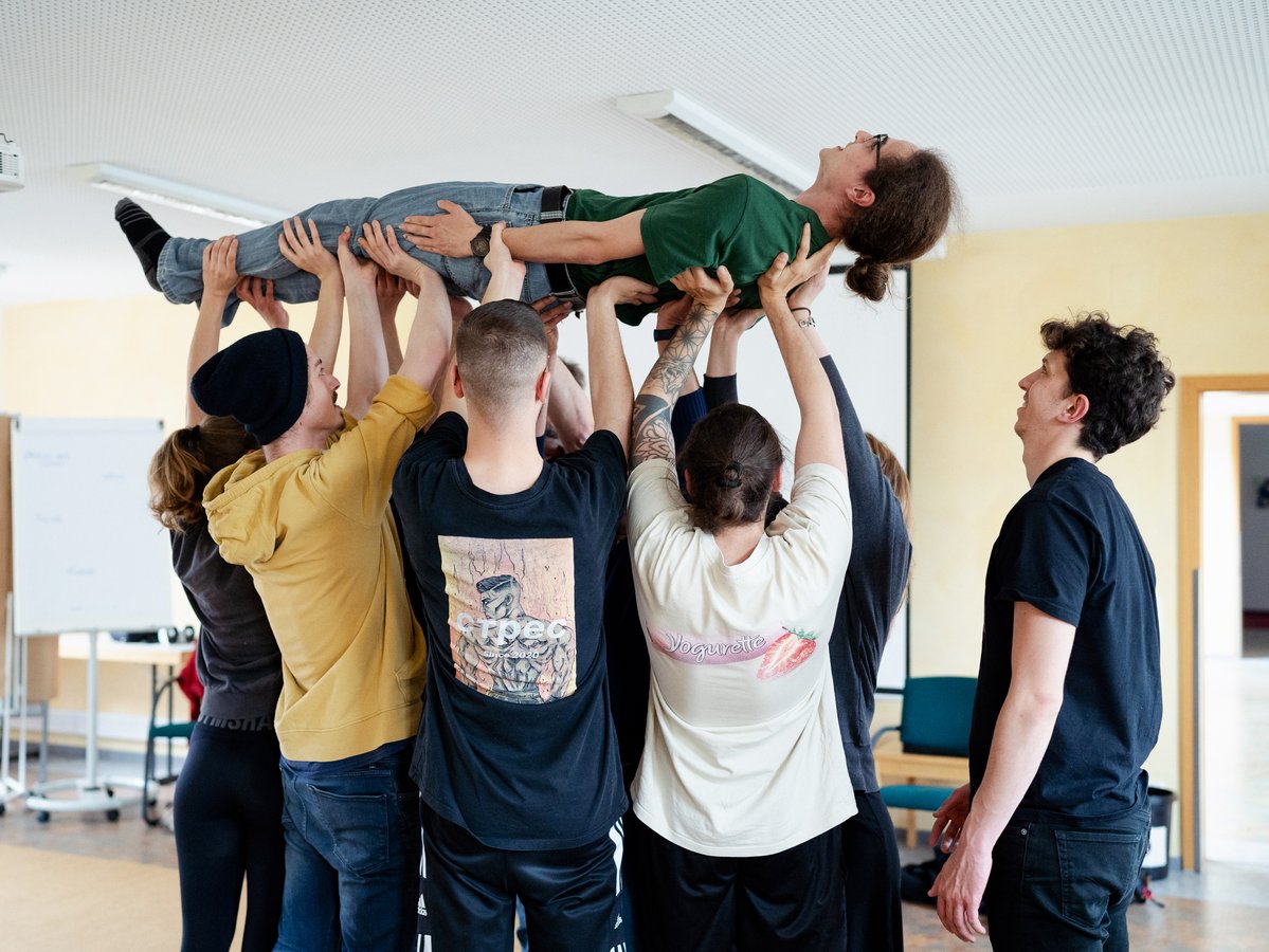 Eine Gruppe hebt eine Person horizontal über den Köpfen in einem Raum, was Teamwork und Vertrauen zeigt.