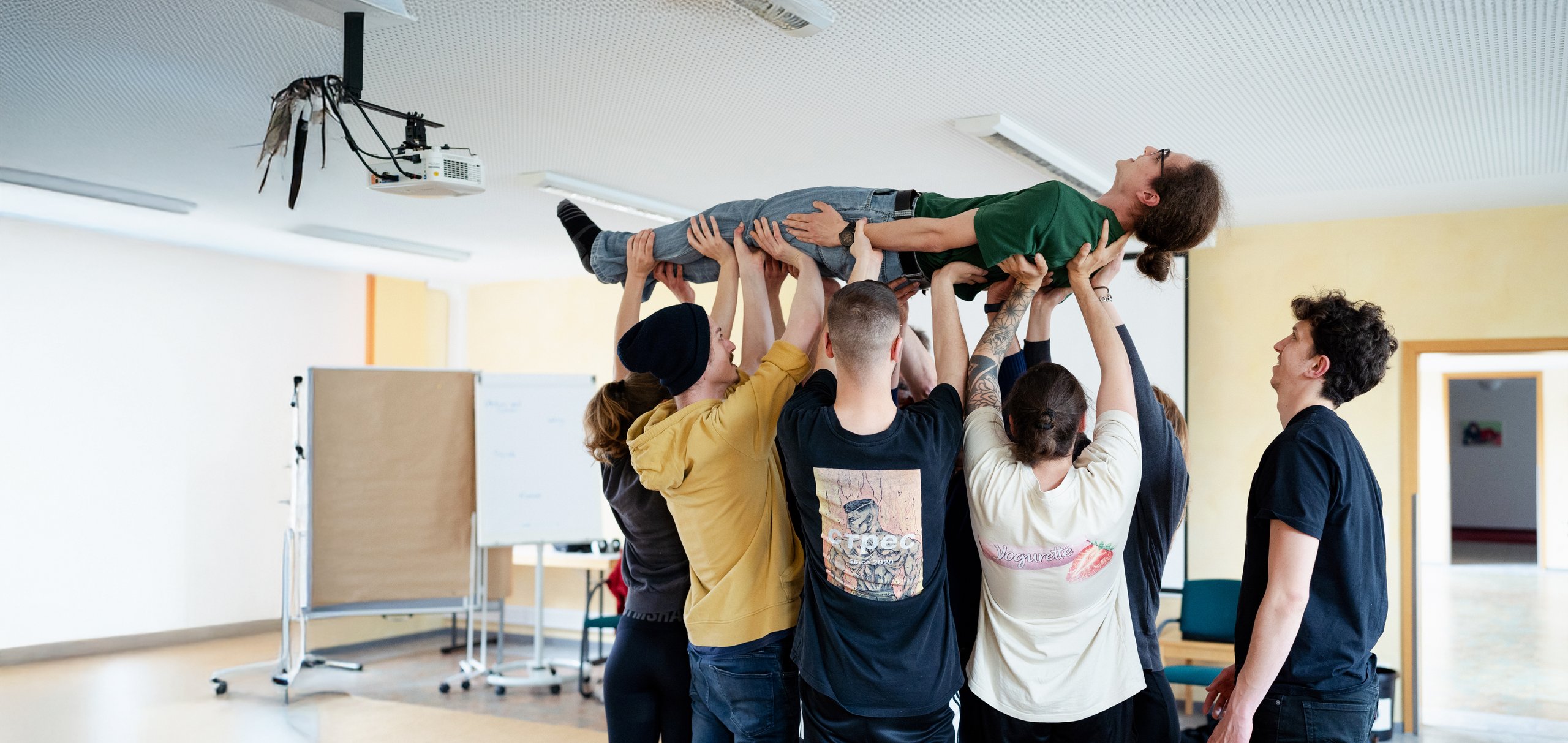 Eine Gruppe hebt eine Person horizontal über den Köpfen in einem Raum, was Teamwork und Vertrauen zeigt.