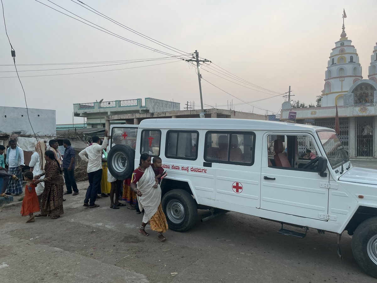 Ein weißes medizinisches Servicefahrzeug mit roten Kreuzsymbolen auf einem Feldweg im ländlichen Indien, umgeben von Menschen.