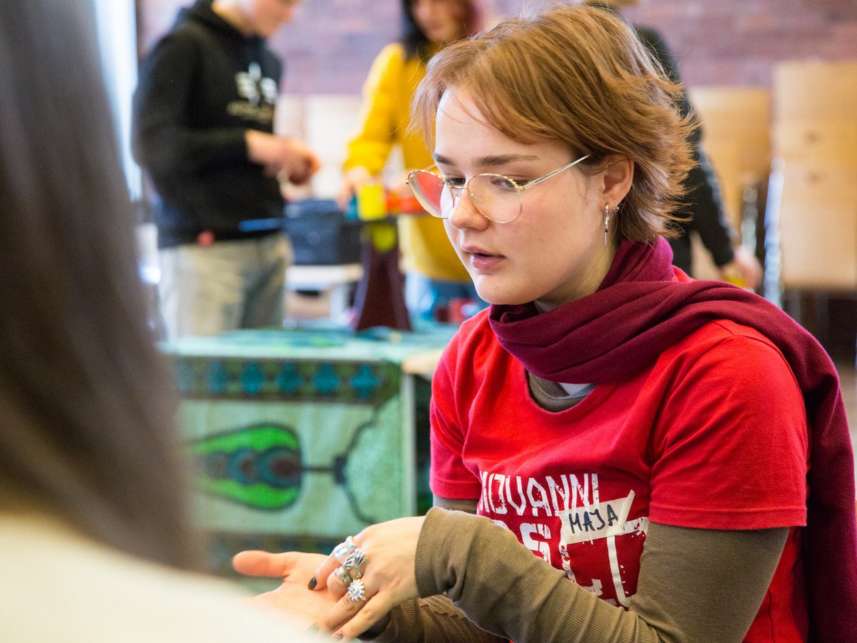 Junge Frau mit Brille und Schal schaut auf ihre Hände, die mit Ringen geschmückt sind, während sie an einer Aktivität teilnimmt.