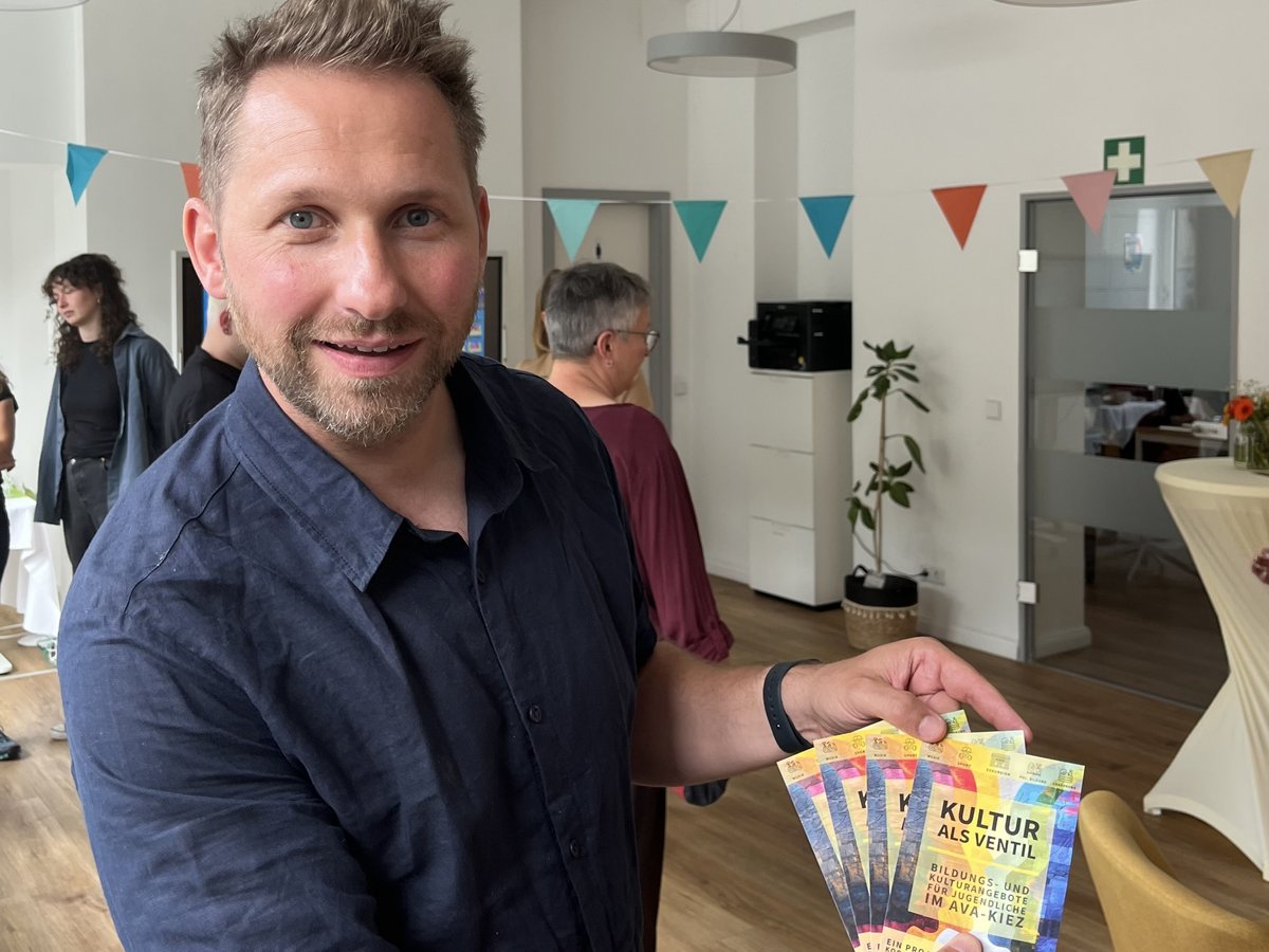 Ein lächelnder Mann im blauen Hemd hält bunte Flyer für 'Kultur als Ventil', ein Bildungs- und Kulturangebot für Jugendliche.