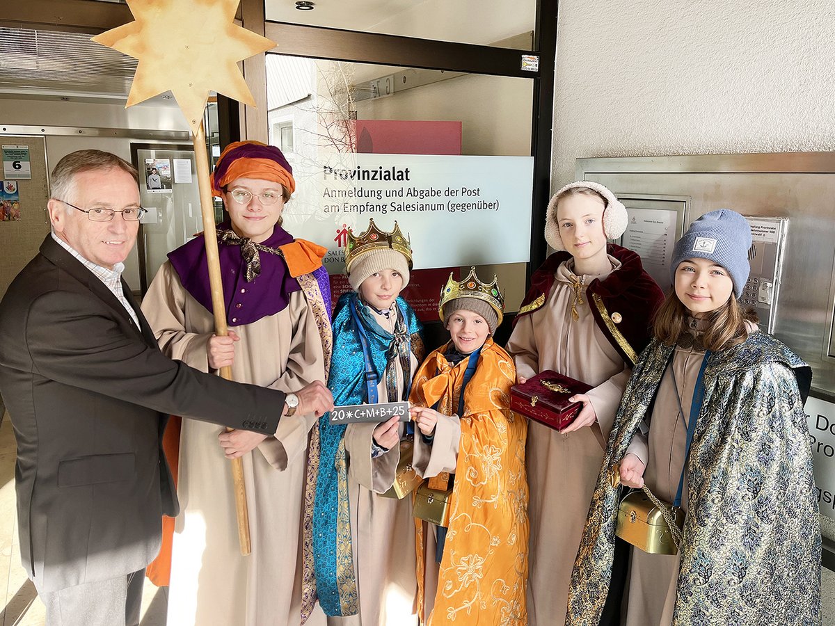 Ein Mann und fünf Sternsingerkinder posieren vor einem Gebäude, ein Kind hält ein Schild mit '20*C+M+B+25'.