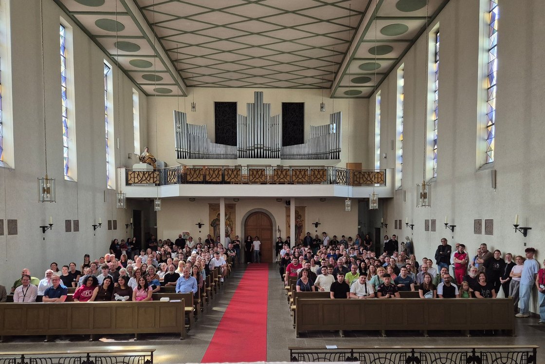 Große Menschengruppe in einer hellen Kirche mit hohen Buntglasfenstern, Holzpfeifenorgel auf der Empore und rotem Teppich.