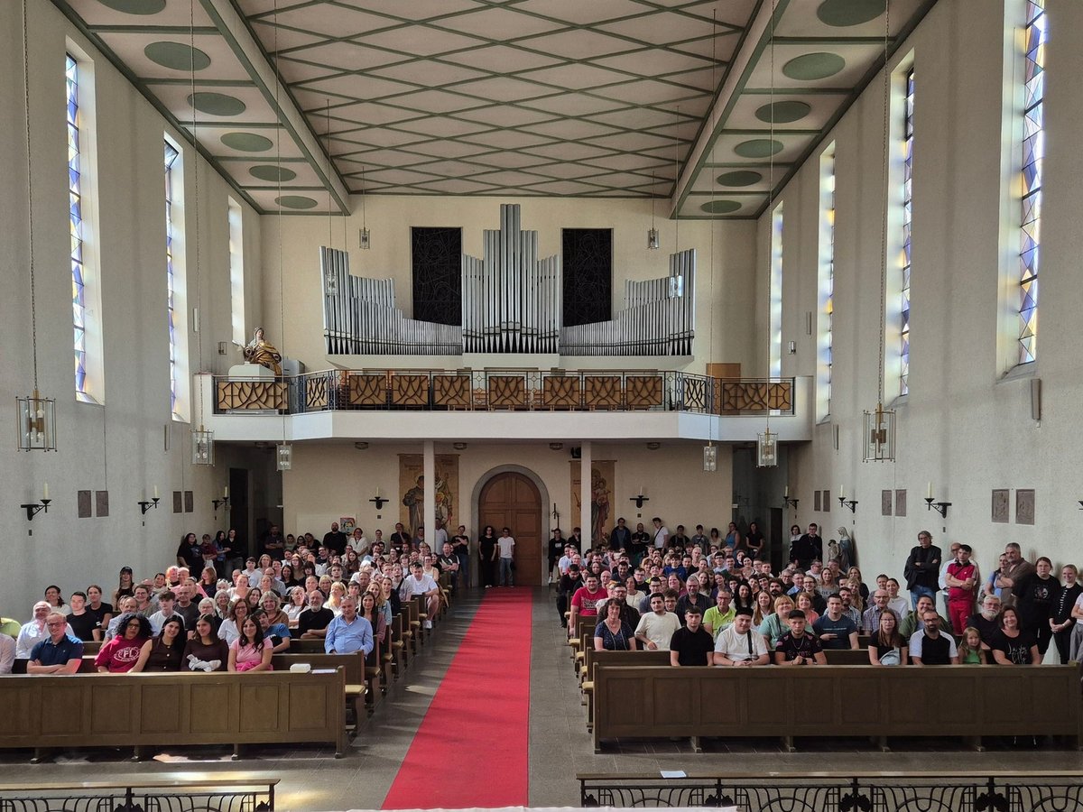 Große Menschengruppe in einer hellen Kirche mit hohen Buntglasfenstern, Holzpfeifenorgel auf der Empore und rotem Teppich.