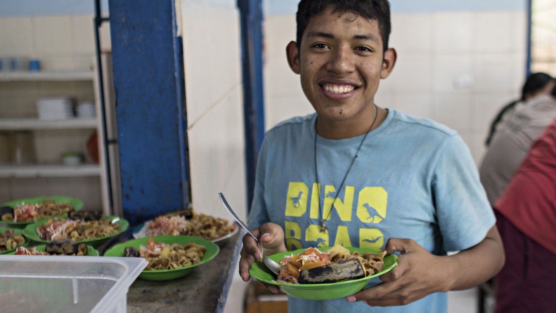 Lächelnder junger Mann im blauen "DINO ROAR" Shirt hält einen grünen Teller mit Essen in einer Mensa.