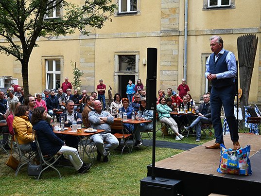 Ein Mann tritt auf einer Außenbühne vor sitzendem Publikum in einem Innenhof auf. Er steht auf einer Tasche vor einem alten Gebäude.