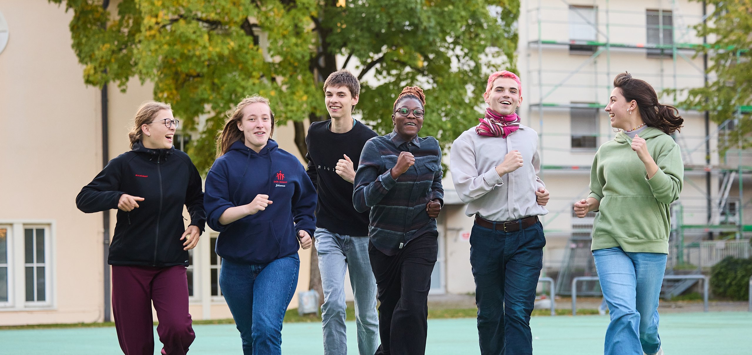 Eine Gruppe lachender junger Menschen rennt auf einem grünen Platz im Freien vor Gebäuden und Bäumen.
