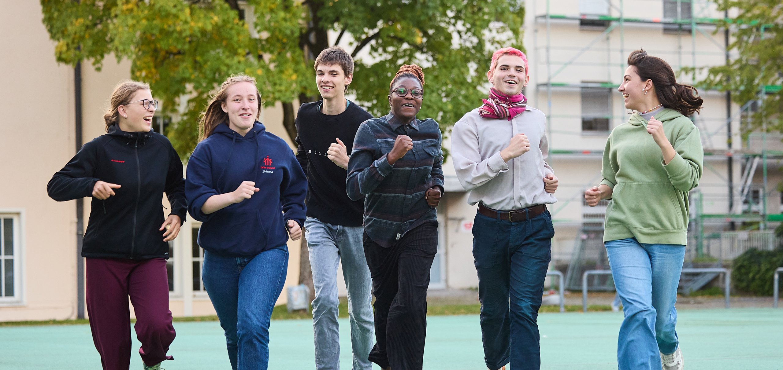 Eine Gruppe lachender junger Menschen rennt auf einem grünen Platz im Freien vor Gebäuden und Bäumen.