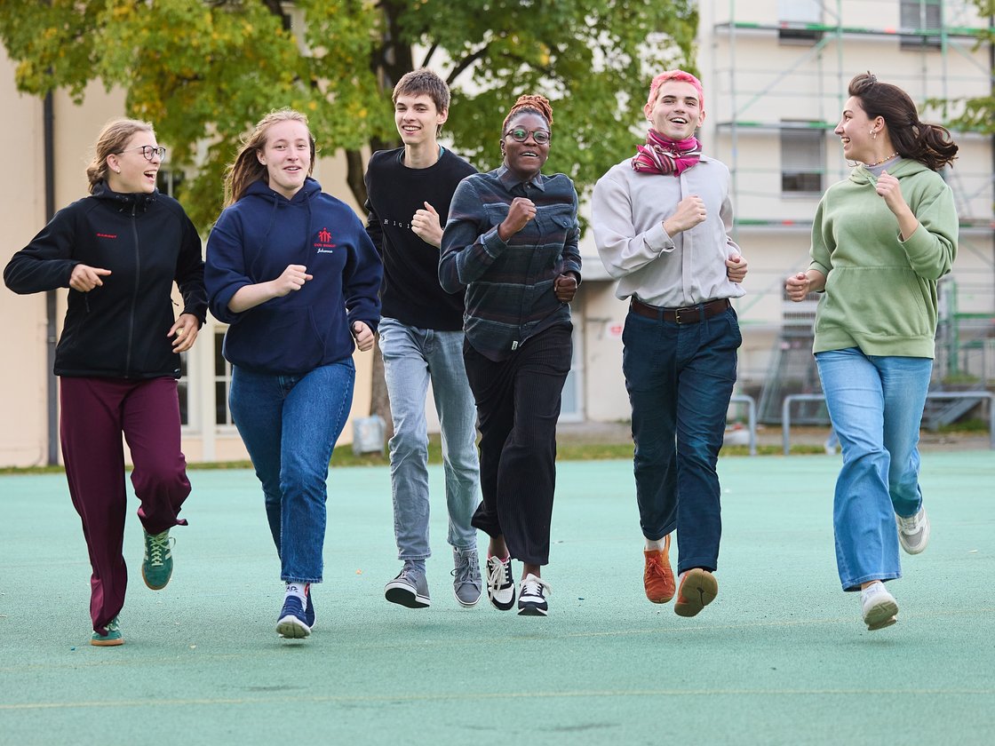 Eine Gruppe lachender junger Menschen rennt auf einem grünen Platz im Freien vor Gebäuden und Bäumen.