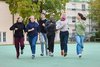 Eine Gruppe lachender junger Menschen rennt auf einem grünen Platz im Freien vor Gebäuden und Bäumen.