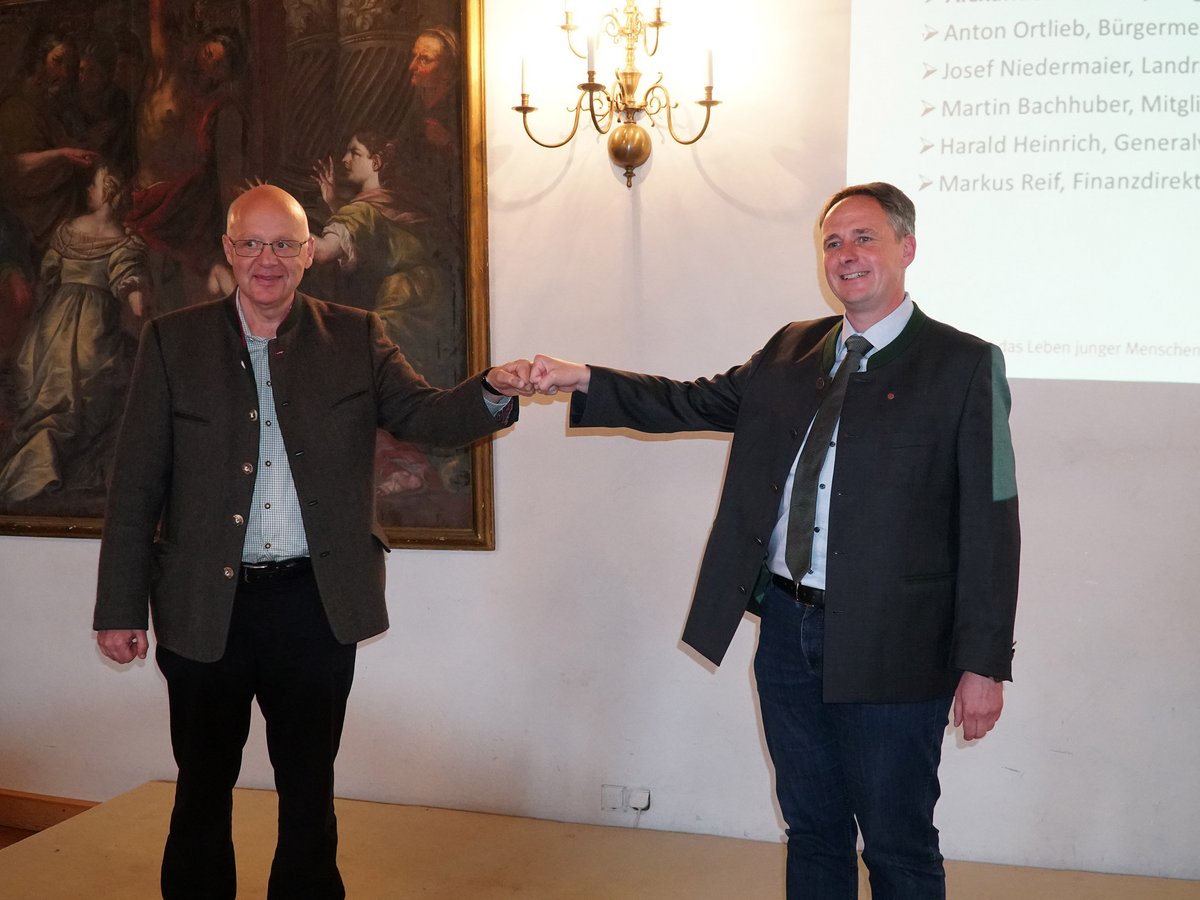 Zwei Männer in Trachtenjacken geben sich einen Faustgruß, im Hintergrund ein Gemälde und eine Projektion mit Namen.