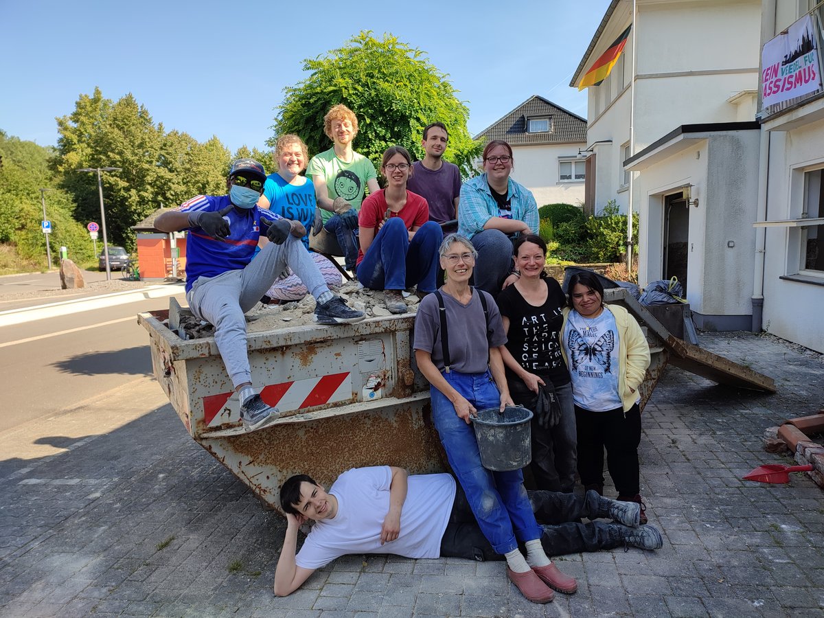 Eine diverse Gruppe von 12 lächelnden Menschen posiert um einen rostigen Container. Im Hintergrund: Haus mit deutscher Flagge und Anti-Rassismus-Banner.