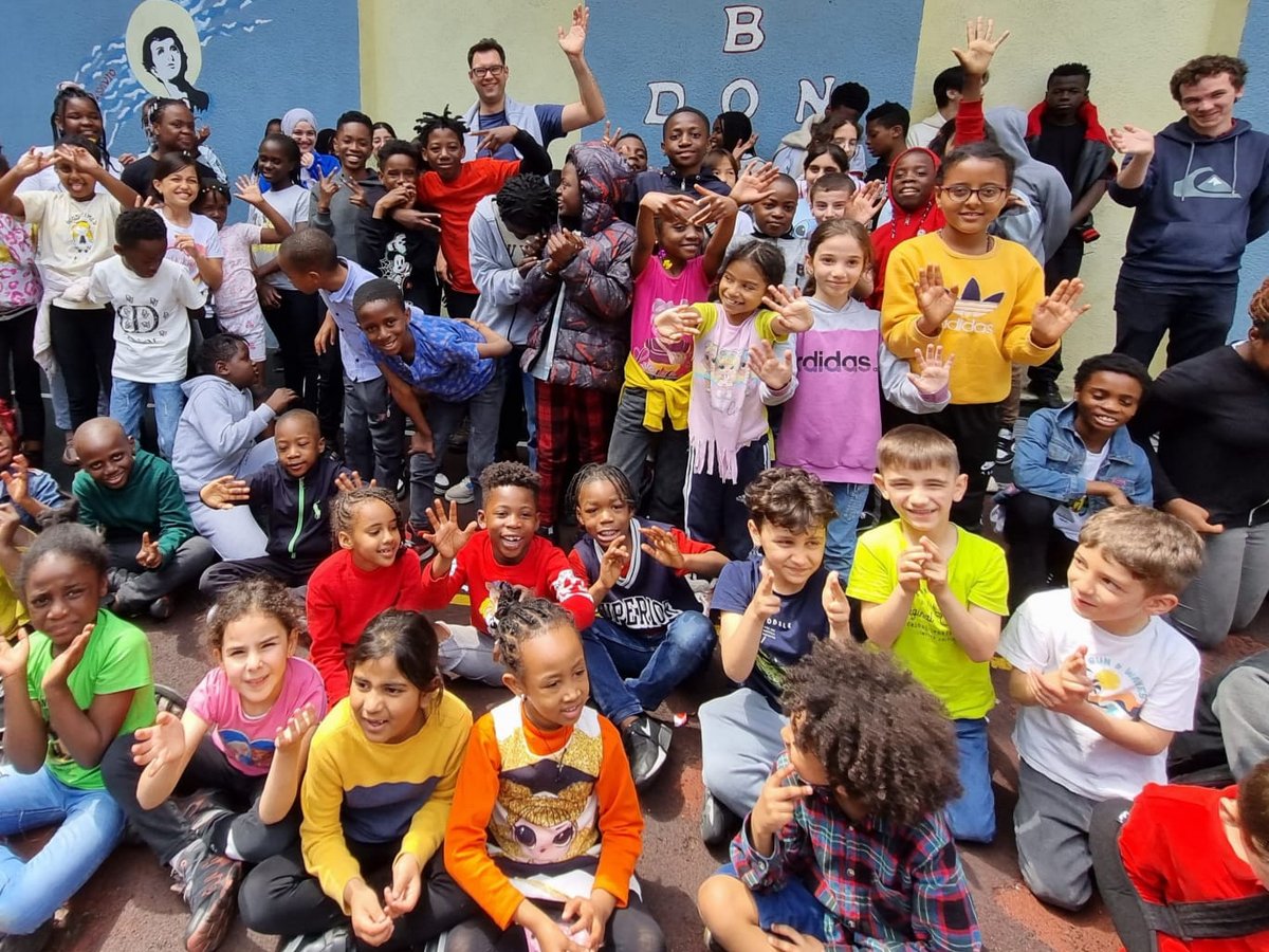 Fröhliche, vielfältige Gruppe von Kindern und Erwachsenen winkt glücklich im Freien vor einer bemalten Wand.