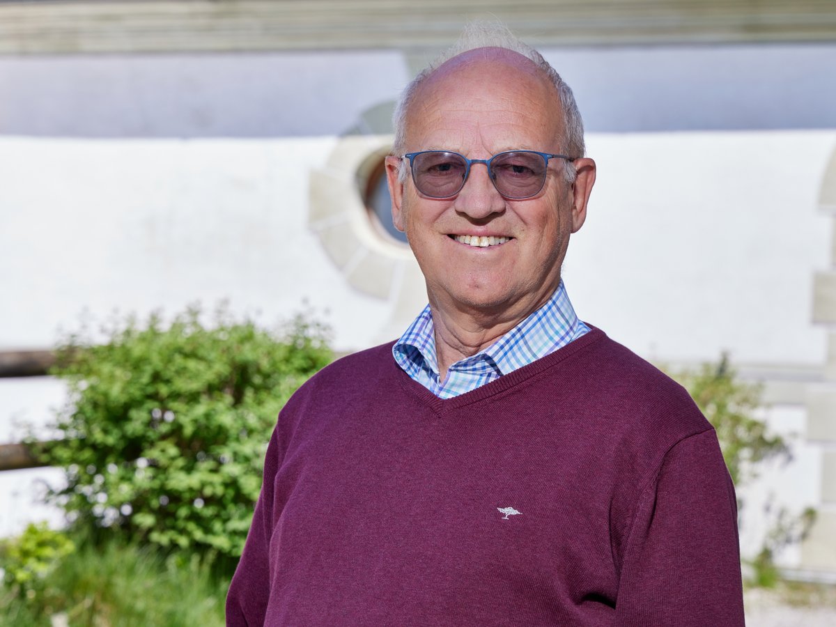 Porträt eines lächelnden älteren Mannes mit Brille und dunkelrotem Pullover vor verschwommenem Hintergrund.