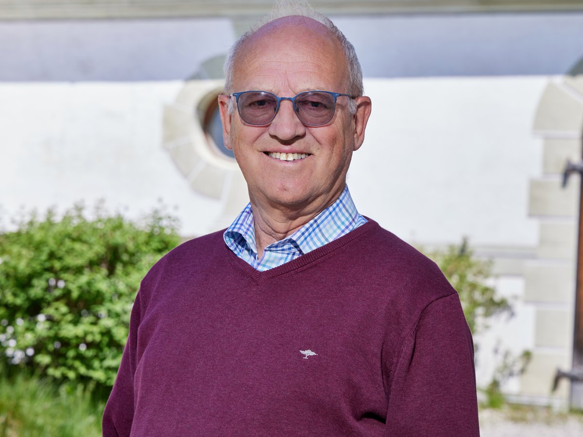 Porträt eines lächelnden älteren Mannes mit Brille und dunkelrotem Pullover vor verschwommenem Hintergrund.