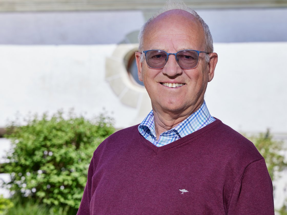 Porträt eines lächelnden älteren Mannes mit Brille und dunkelrotem Pullover vor verschwommenem Hintergrund.