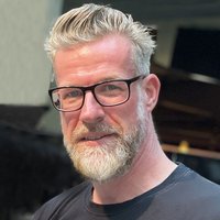 Porträt eines lächelnden Mannes mit grauem Bart, schwarzer Brille und gestyltem grauem Haar in einem schwarzen T-Shirt.