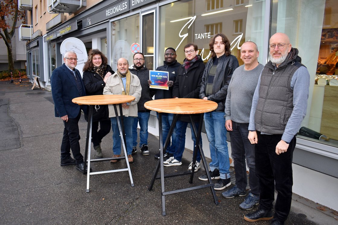 Neun lächelnde Personen, Männer und Frauen, stehen vor einem Geschäft. Einer hält ein Schild: 'Wir bilden aus 2025'.