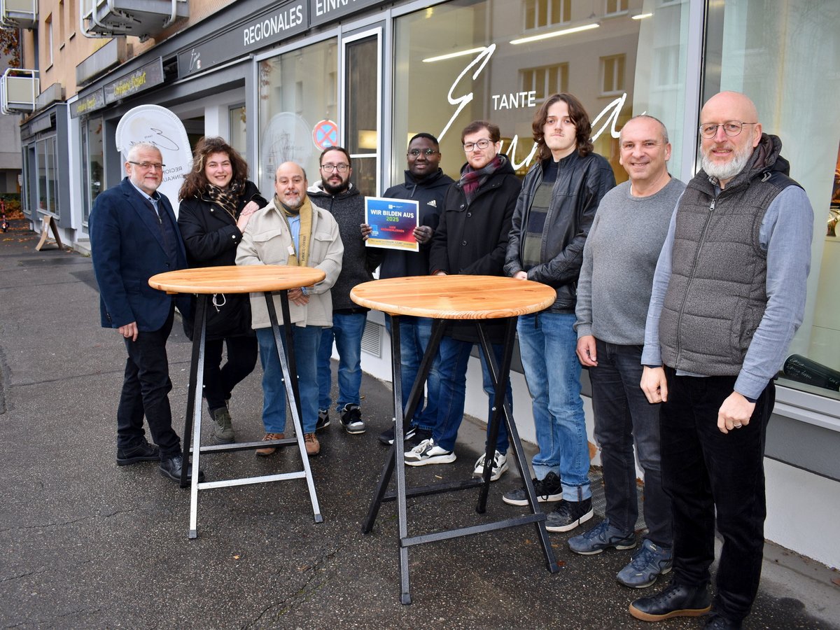 Neun lächelnde Personen, Männer und Frauen, stehen vor einem Geschäft. Einer hält ein Schild: 'Wir bilden aus 2025'.