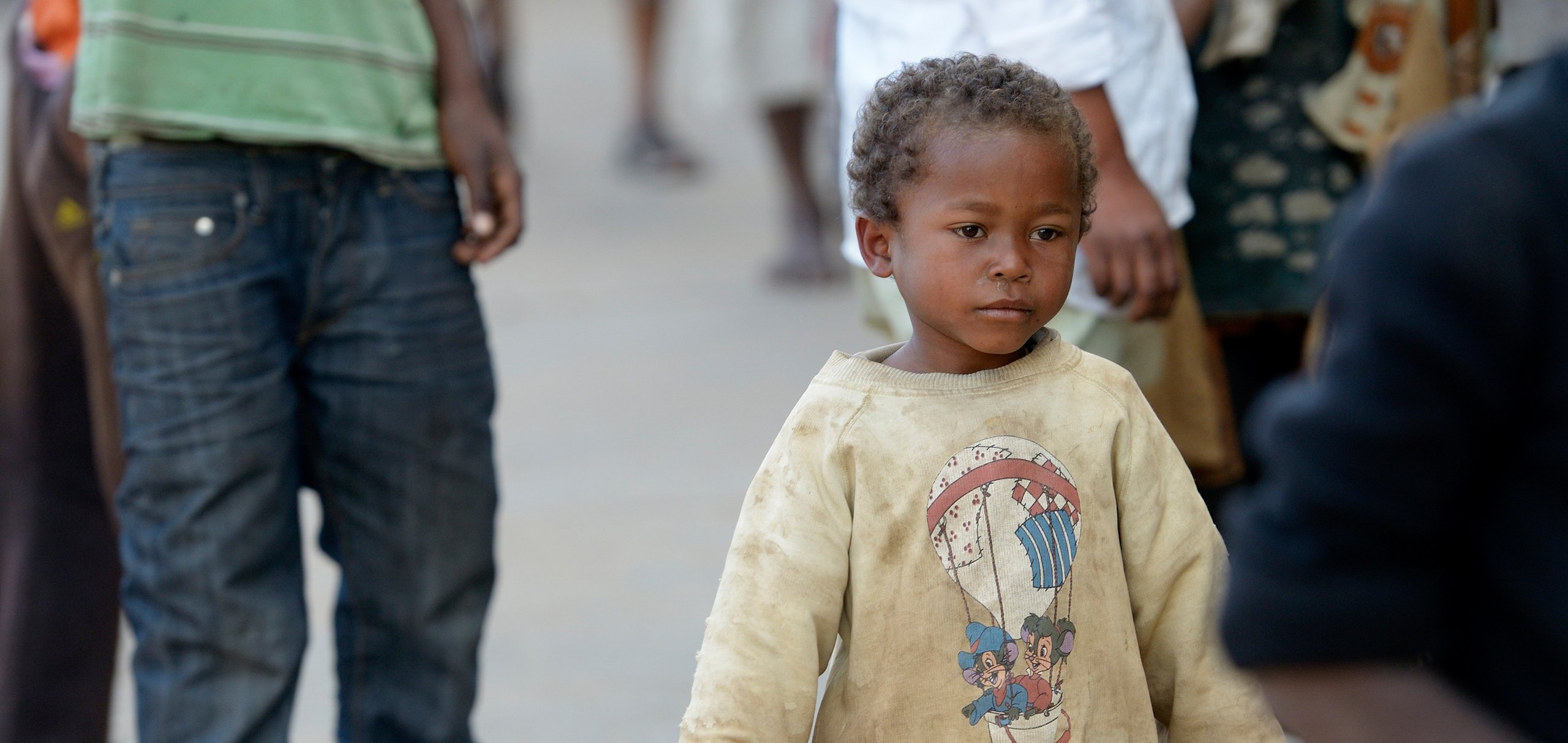 Ein kleiner Junge mit lockigem Haar und schmutzigem Pullover mit Heißluftballon-Motiv blickt nach rechts in belebter Umgebung.