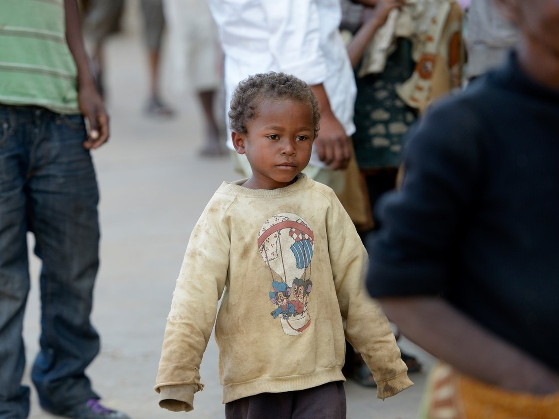 Ein kleiner Junge mit lockigem Haar und schmutzigem Pullover mit Heißluftballon-Motiv blickt nach rechts in belebter Umgebung.