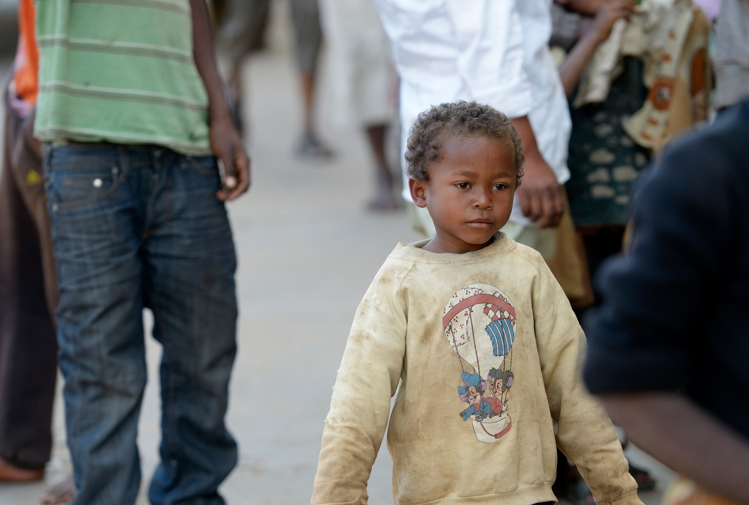 Ein kleiner Junge mit lockigem Haar und schmutzigem Pullover mit Heißluftballon-Motiv blickt nach rechts in belebter Umgebung.